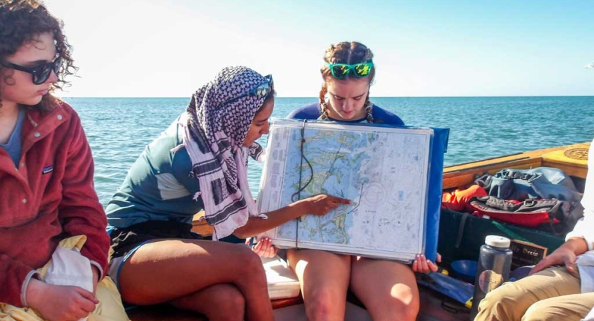 Three people sitting on a sailboat examine a map. Behind them the blue ocean expands to the horizon. 